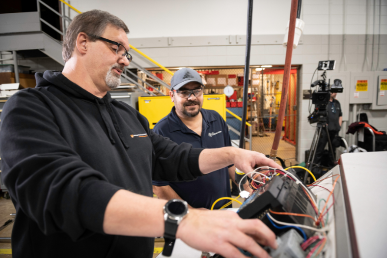 An instructor and student, both in safety glasses are gathered around a circuit board.